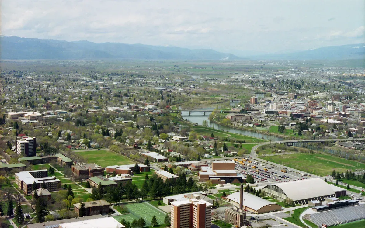 An aerial view of Missoula, Montana, with the Clark Fork River winding through downtown and the University of Montana campus in the foreground, framed by mountains