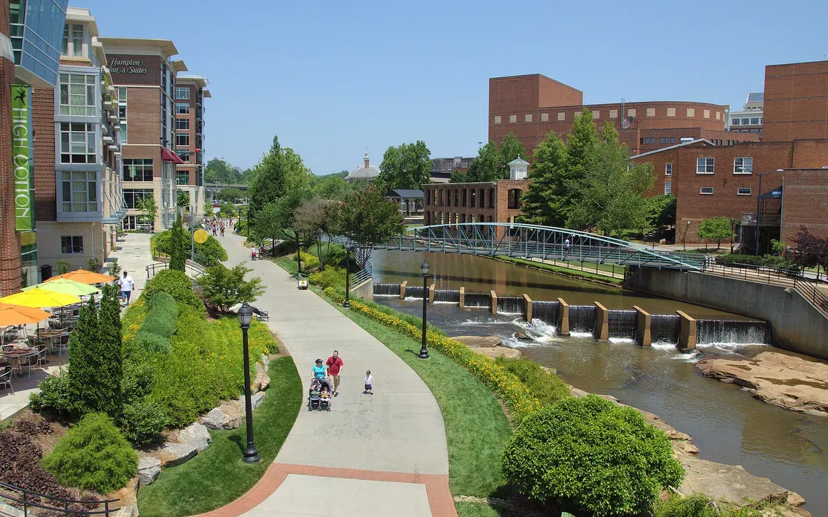 A riverside walking path along the Reedy River in downtown Greenville, South Carolina, with a pedestrian bridge, small waterfalls, and brick buildings on a sunny day